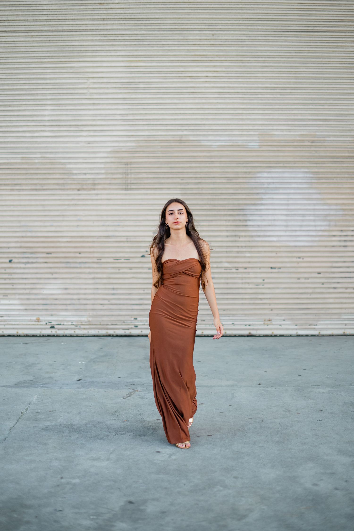 Woman in rust brown strapless maxi dress posing against corrugated metal wall on gray concrete.