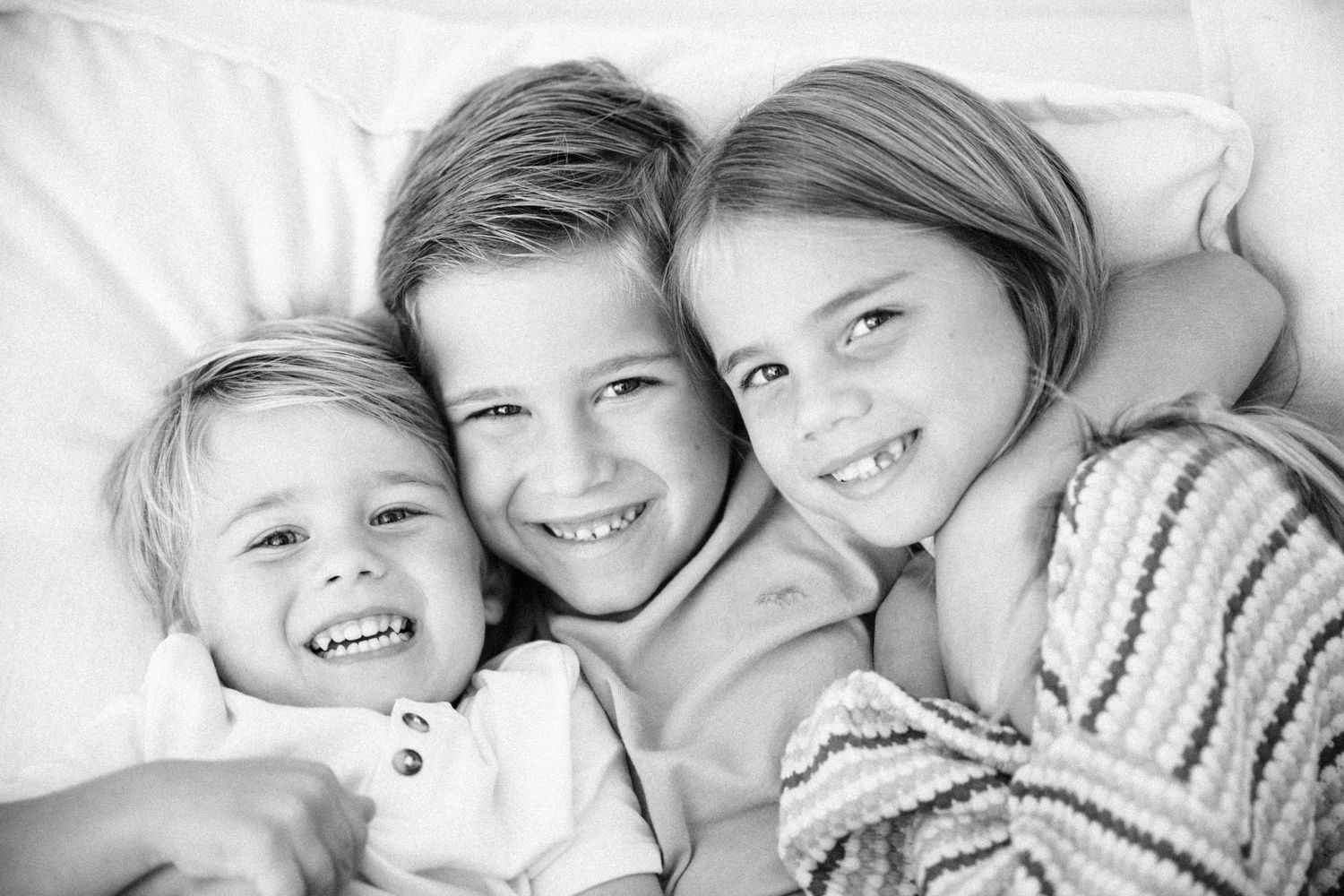 Black and white photo of three happy children smiling and cuddling together on a white couch.