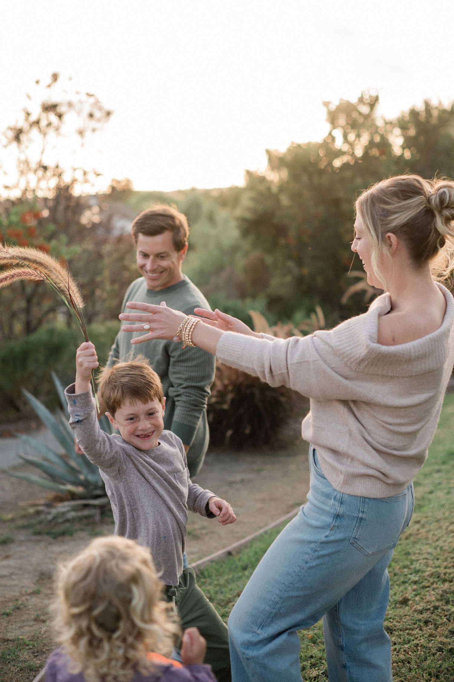 Family playing outdoors at sunset, with a laughing child holding grass reeds between smiling parents in a garden.