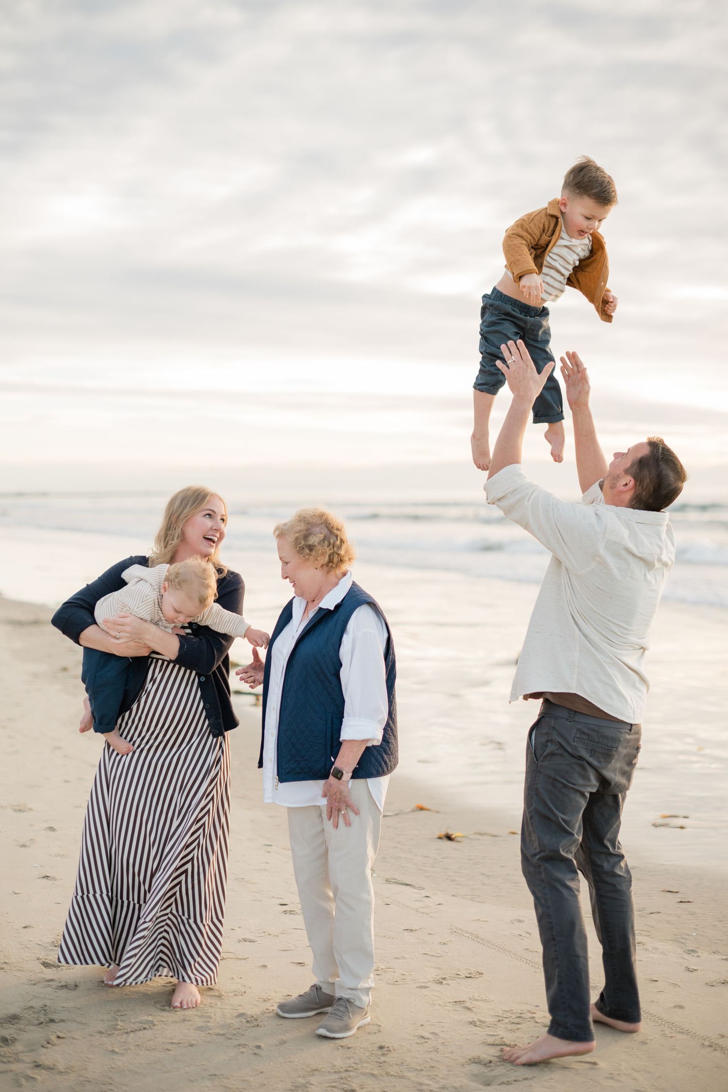 Family enjoys beach fun as dad tosses toddler in air while mom and grandma watch and laugh.