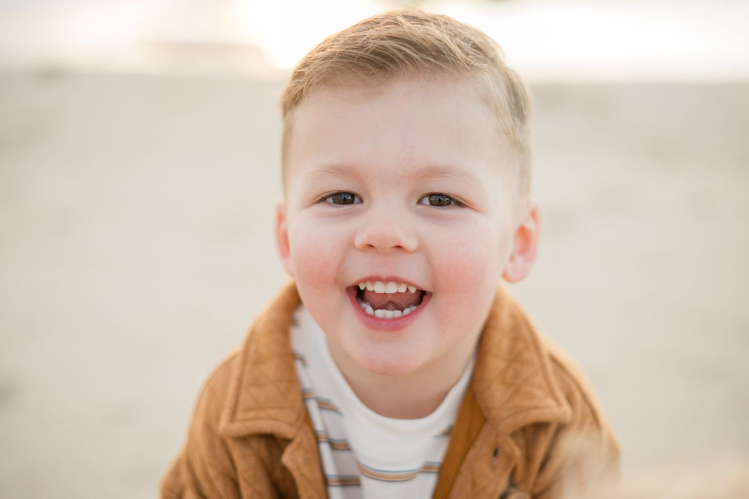 A happy toddler boy with short blond hair smiling outdoors, wearing a brown jacket over a striped shirt.