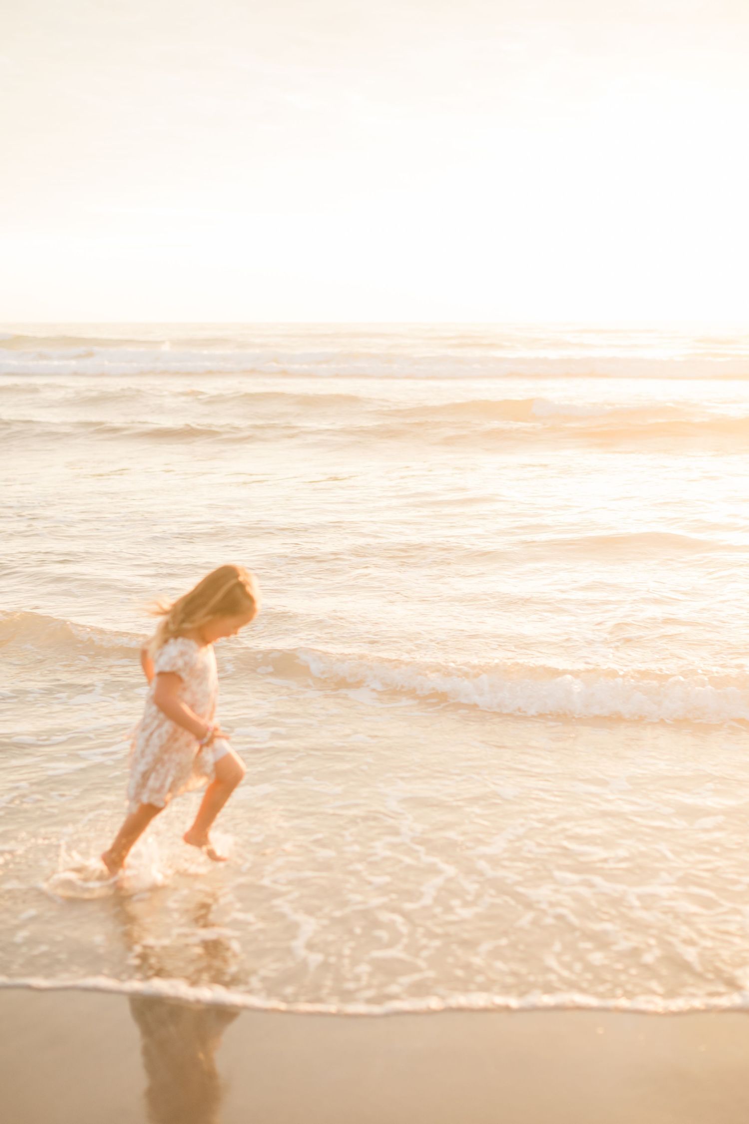 A young girl in a white dress runs through shallow ocean waves on a golden sunlit beach.