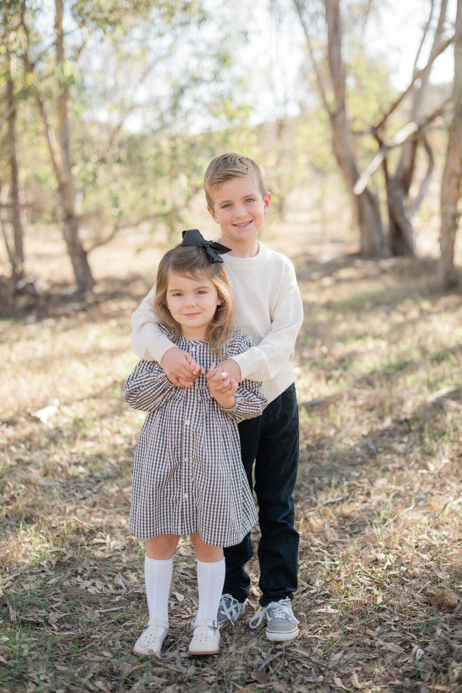 Two young siblings pose outdoors in a wooded area, girl in plaid dress and boy in cream sweater with black pants.