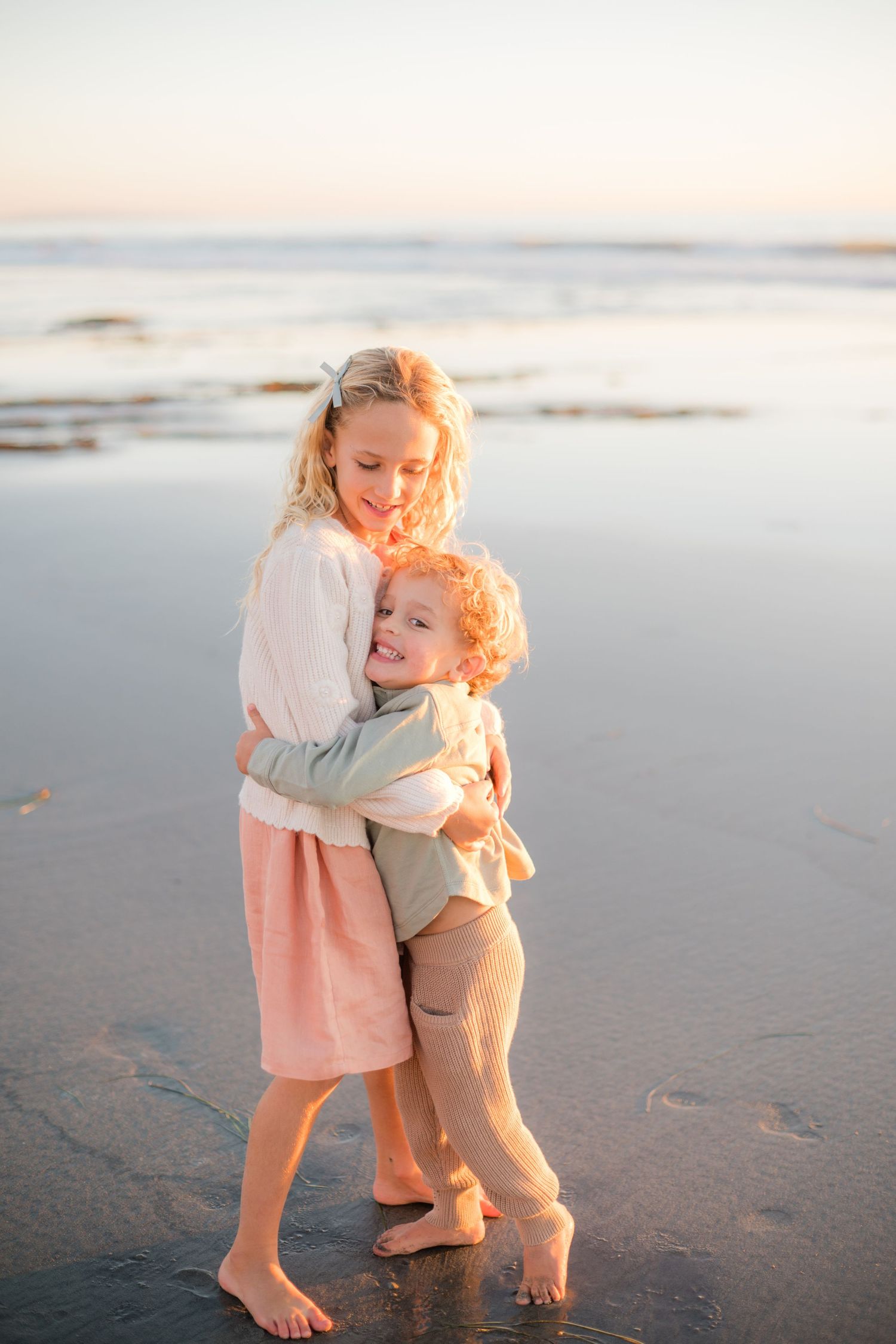 Two young siblings share a warm hug on a sandy beach at golden sunset.