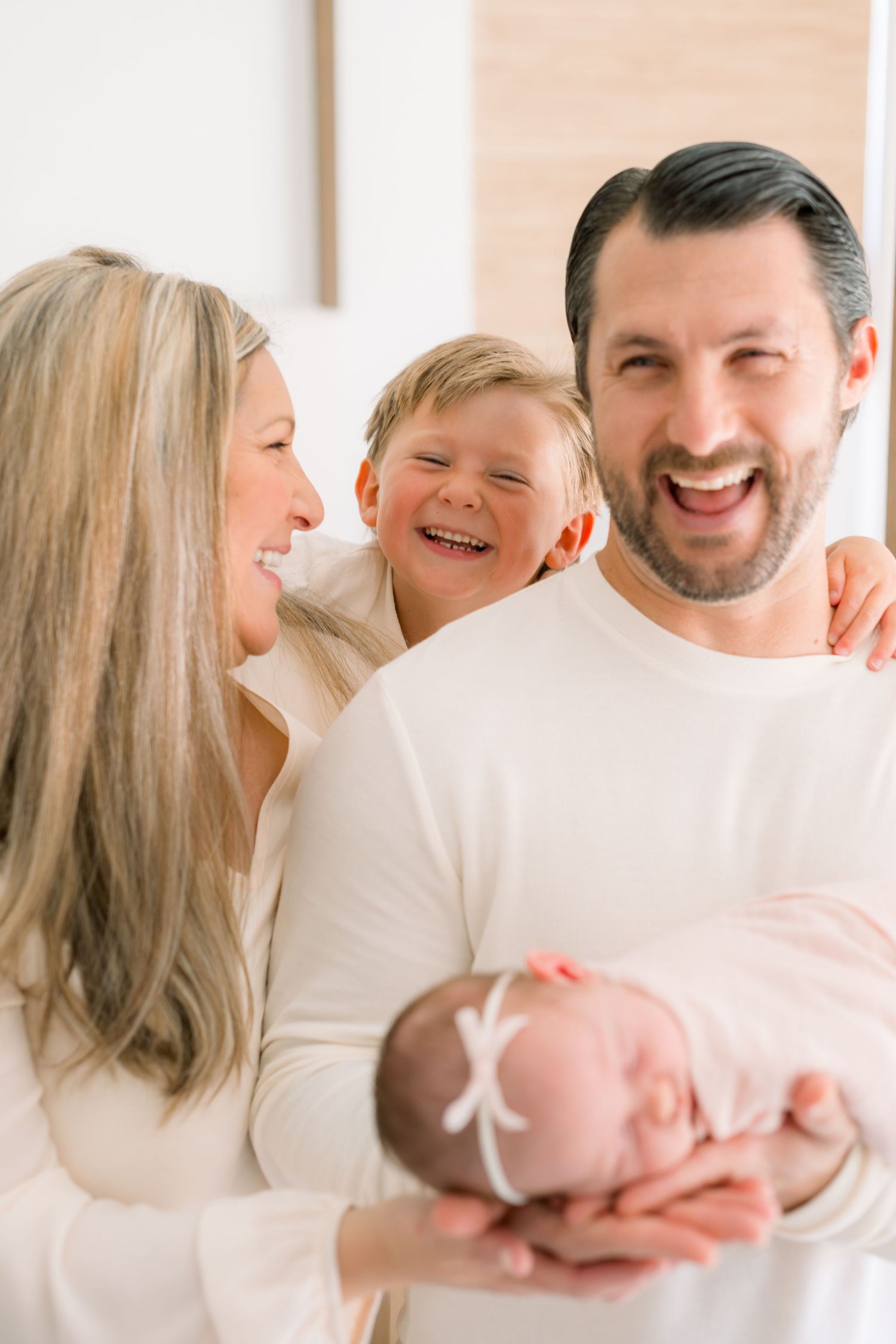 Happy family with parents, toddler boy, and newborn baby girl in white outfits sharing a joyful moment together.