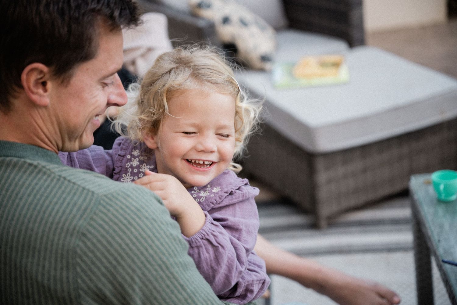 A father laughing with his young blonde toddler girl on an outdoor patio, sharing a joyful moment together.