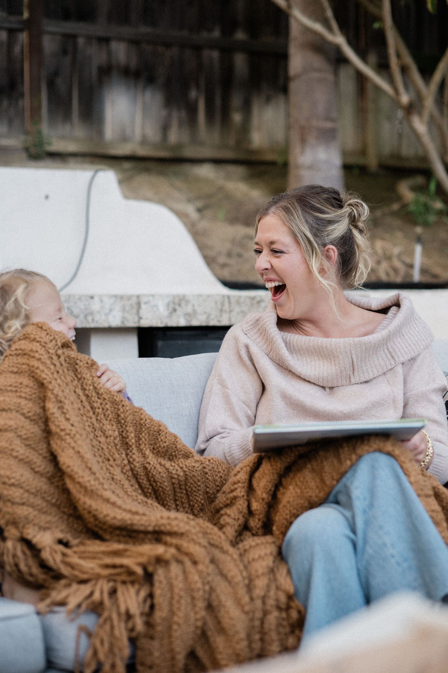 A laughing blonde woman in beige sweater and jeans sits outdoors with child under brown knit blanket holding tablet.