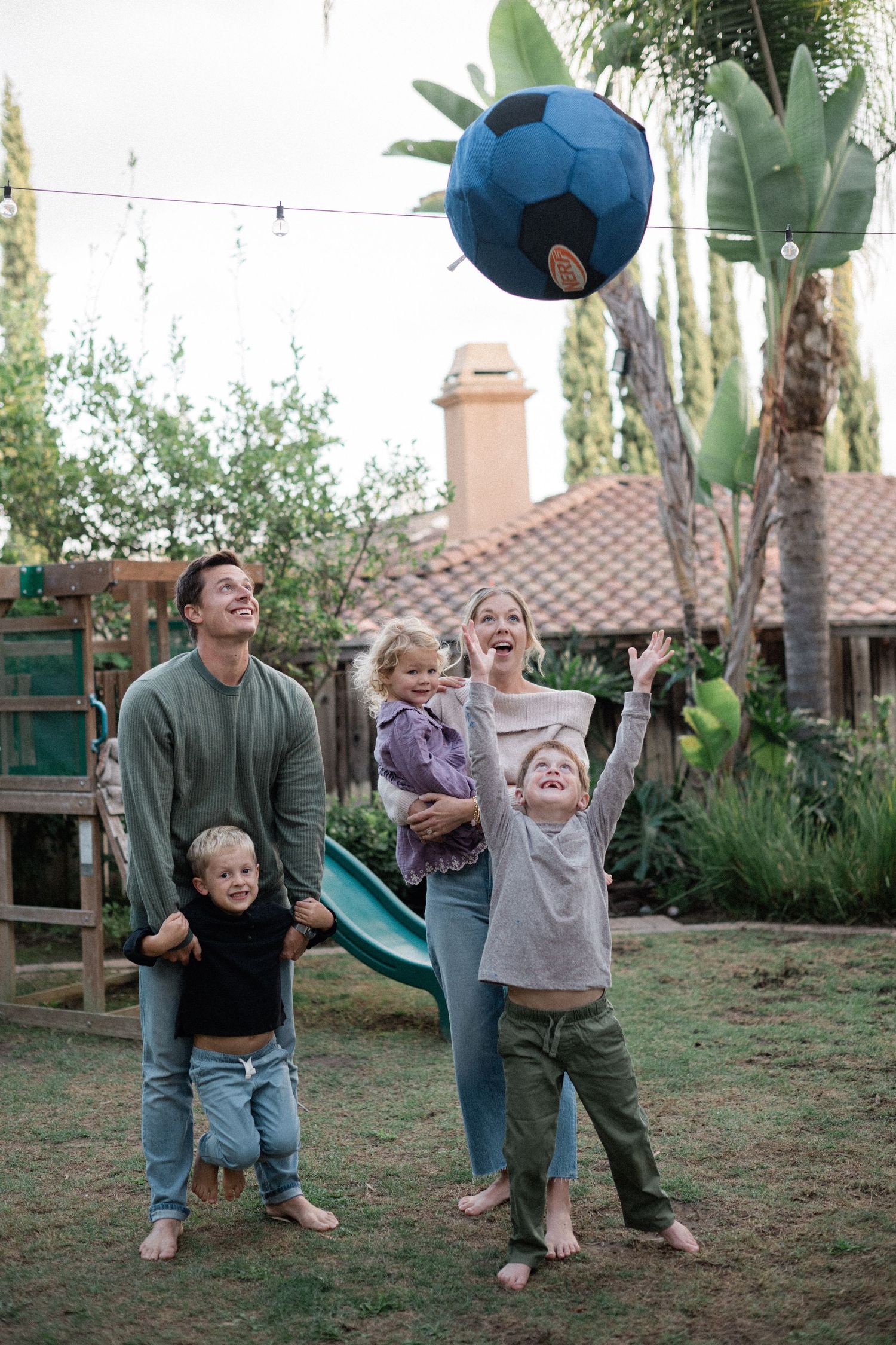 Happy family playing with a soccer ball in a backyard garden, with two parents and three young children.