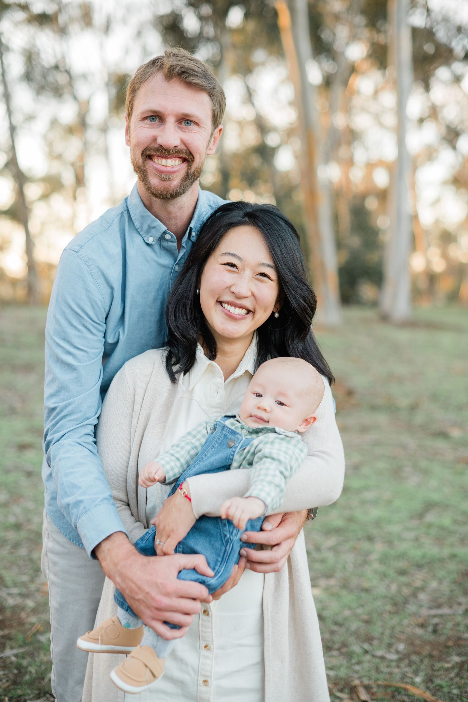 Happy family portrait of smiling couple holding their baby outdoors in a sunlit park setting.