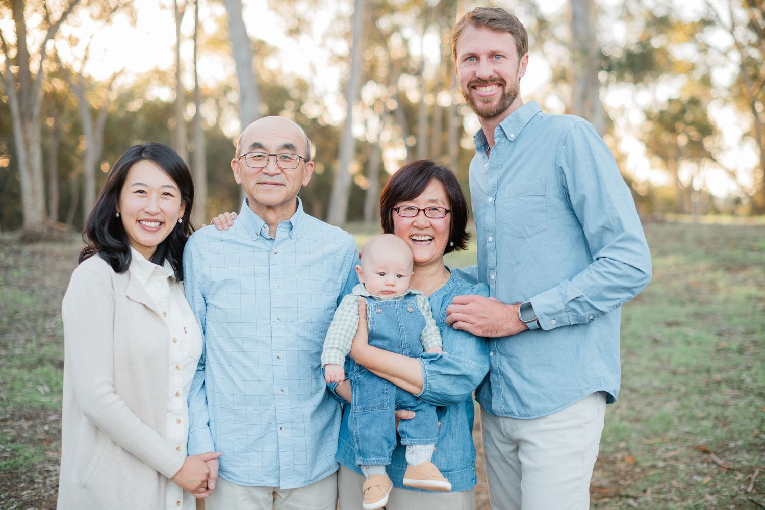 Multi-generational family of five smiling together outdoors in a sunny park setting.