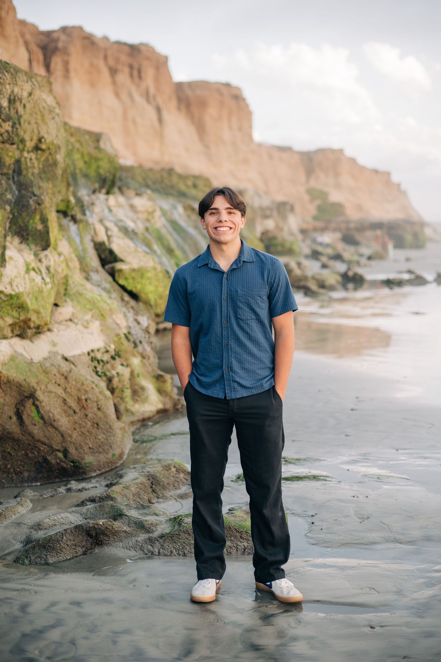 Smiling teen boy in blue shirt and black pants standing on rocky beach with coastal cliffs behind him.