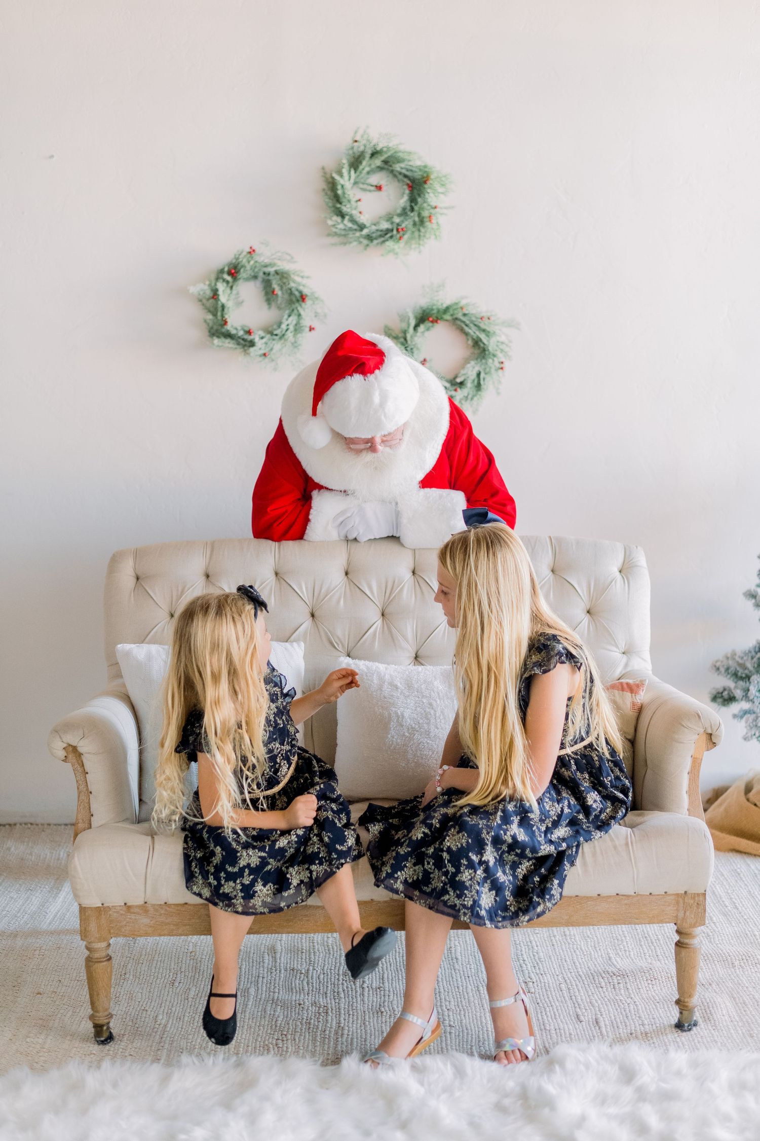 Two blonde girls in floral dresses sit with Santa Claus on a cream tufted sofa, looking at a gift together.