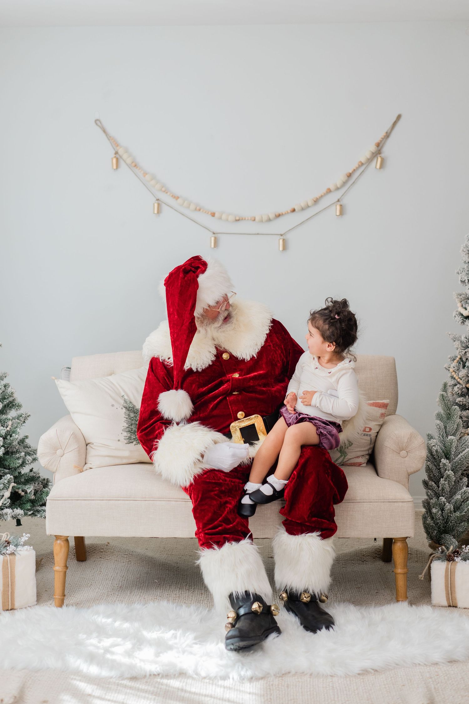 A young girl sits on Santa Claus's lap on a cream sofa surrounded by Christmas trees and gifts.