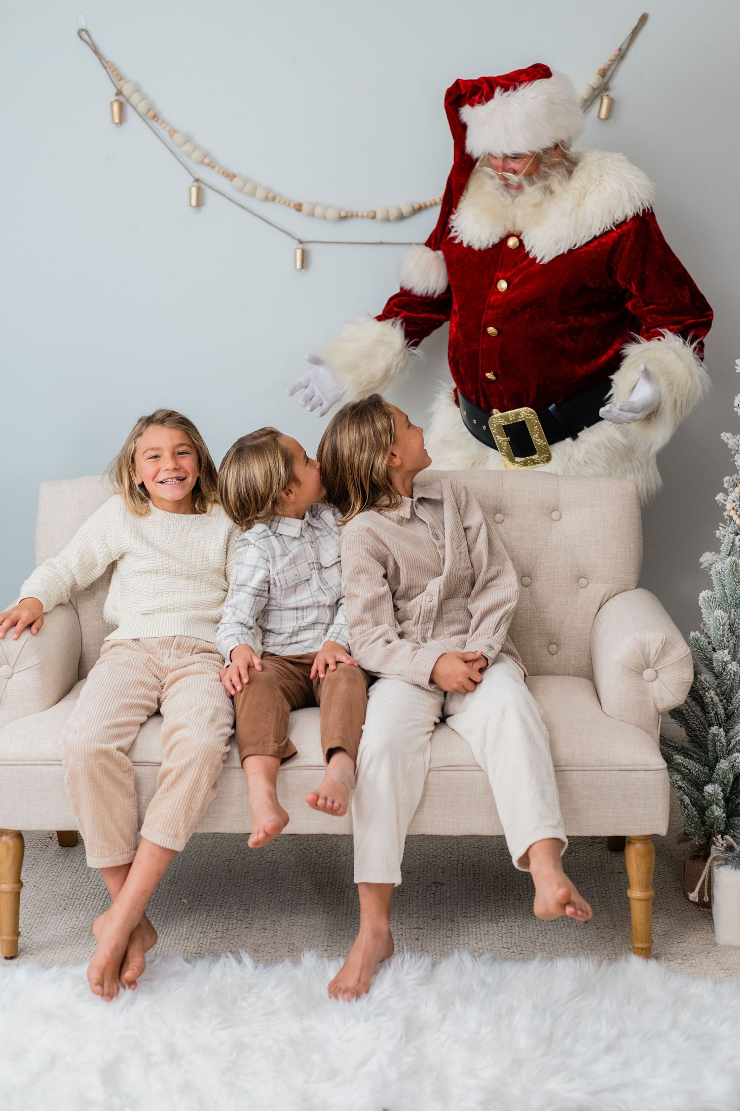 Three happy barefoot children sit on a cream sofa laughing with Santa Claus in a festive decorated room.