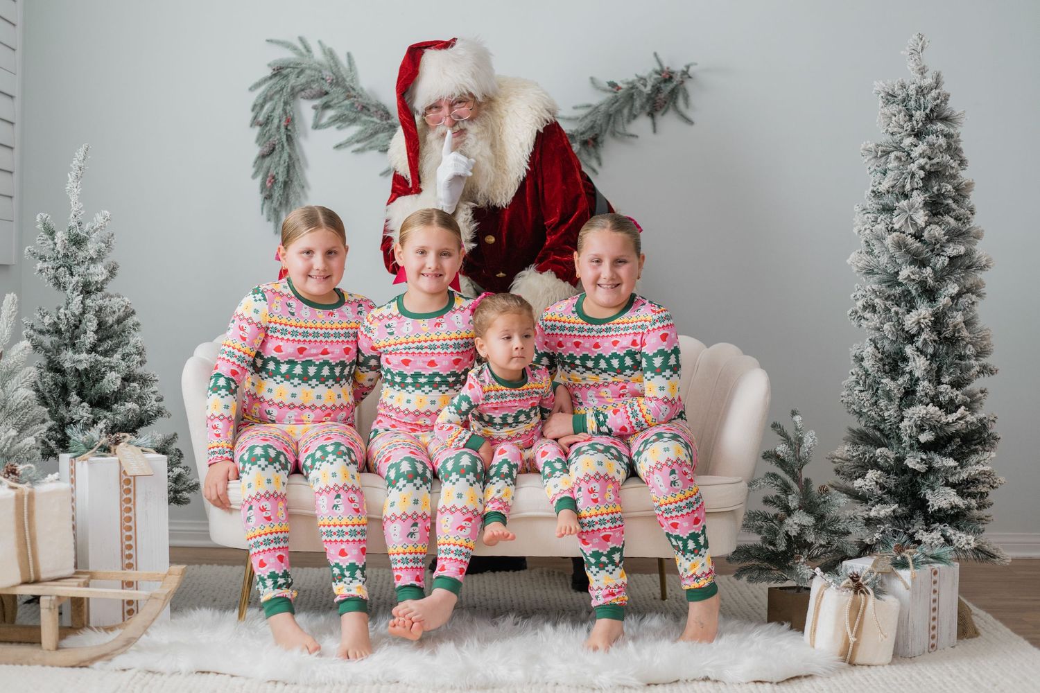 Four children in matching Christmas pajamas pose with Santa Claus among snowy trees and gifts.