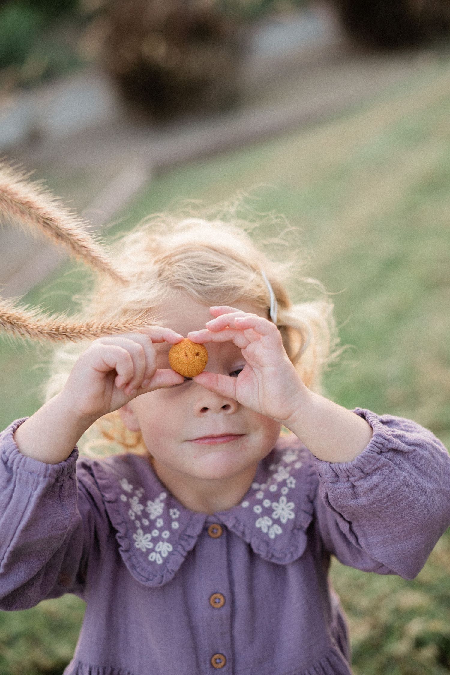Blonde toddler girl in purple floral dress covering eyes with small orange fruit.