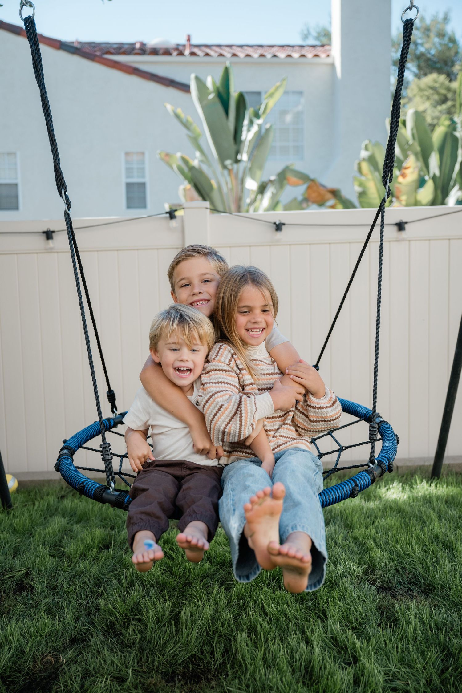 Three happy barefoot children laughing and hugging on a blue rope swing in a sunny backyard.