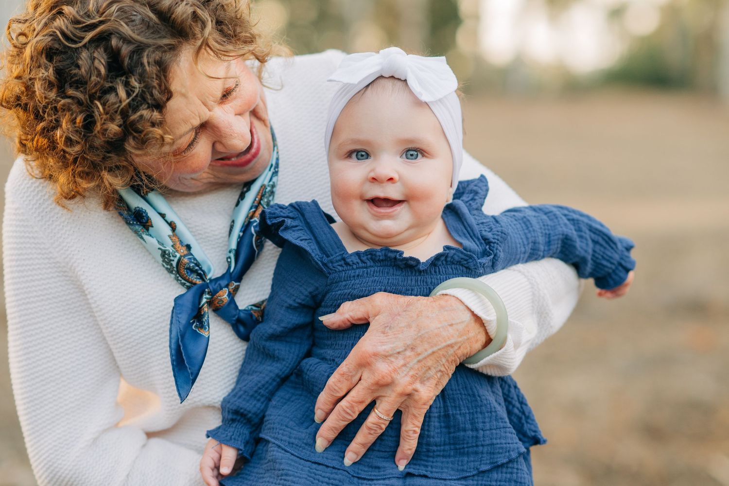 Grandmother with curly hair holds smiling baby girl in blue dress and white bow headband outdoors.