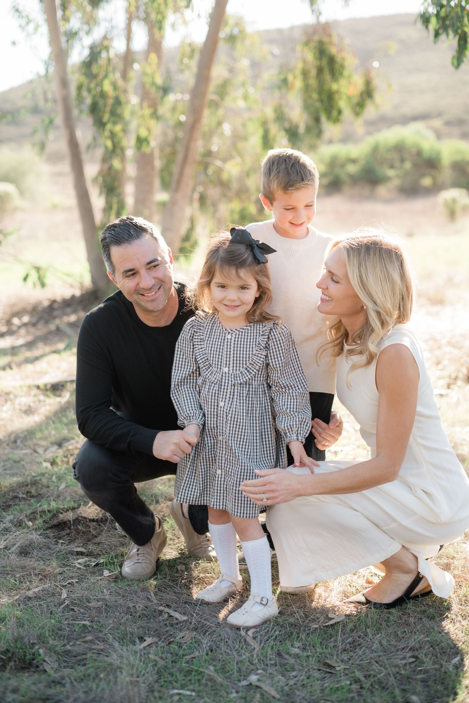 Happy family of four posing outdoors in natural setting with trees, parents crouching beside young children in coordinated outfits.