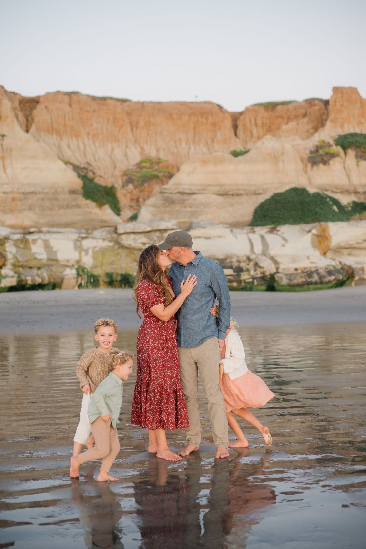 A couple shares a kiss on a wet beach at sunset while their three children play around them.