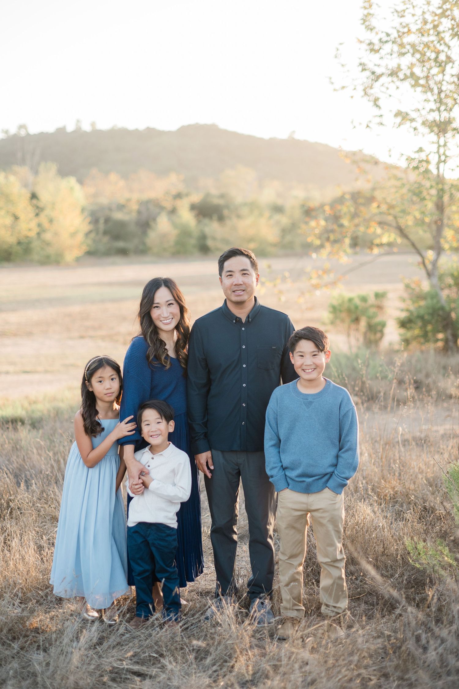Family of five posing outdoors in a golden field at sunset, parents with three children in coordinated blue outfits.