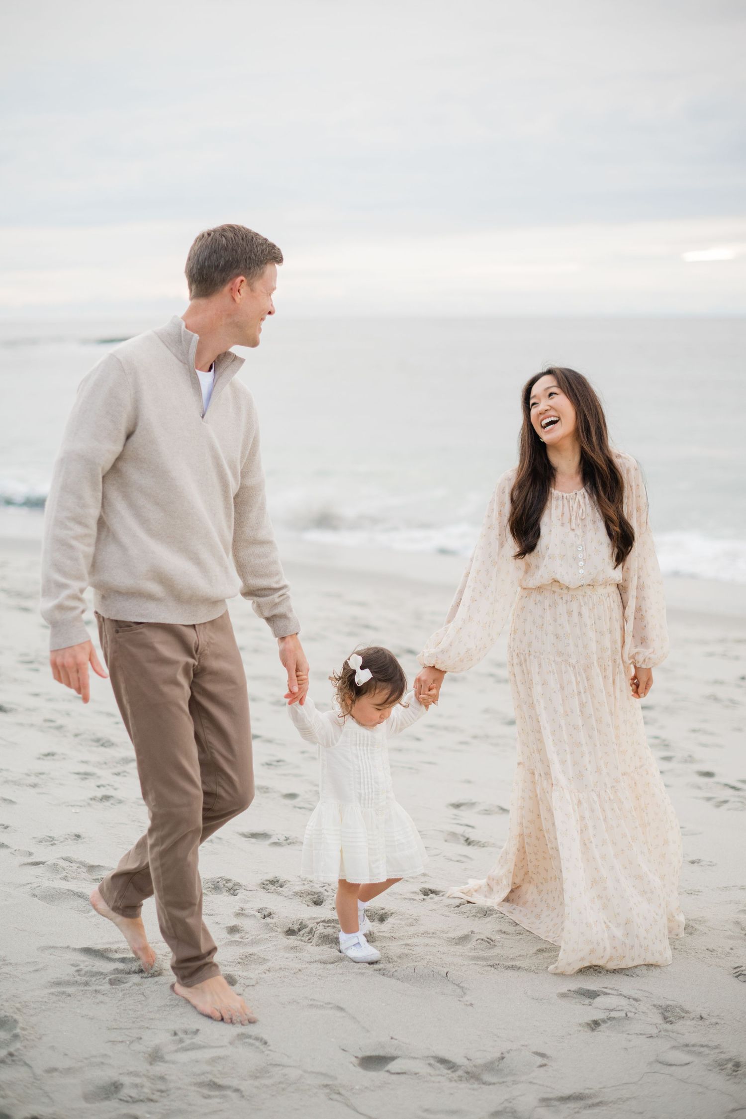 Happy family of three walking hand-in-hand on a sandy beach, parents swinging their toddler.