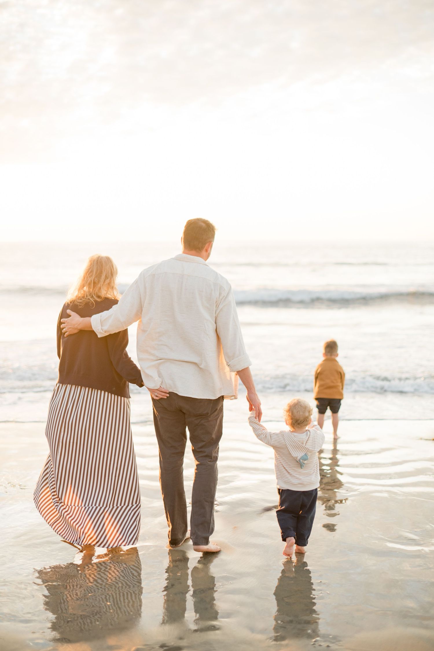 Family walking toward ocean waves at golden hour beach, parents with two young children.