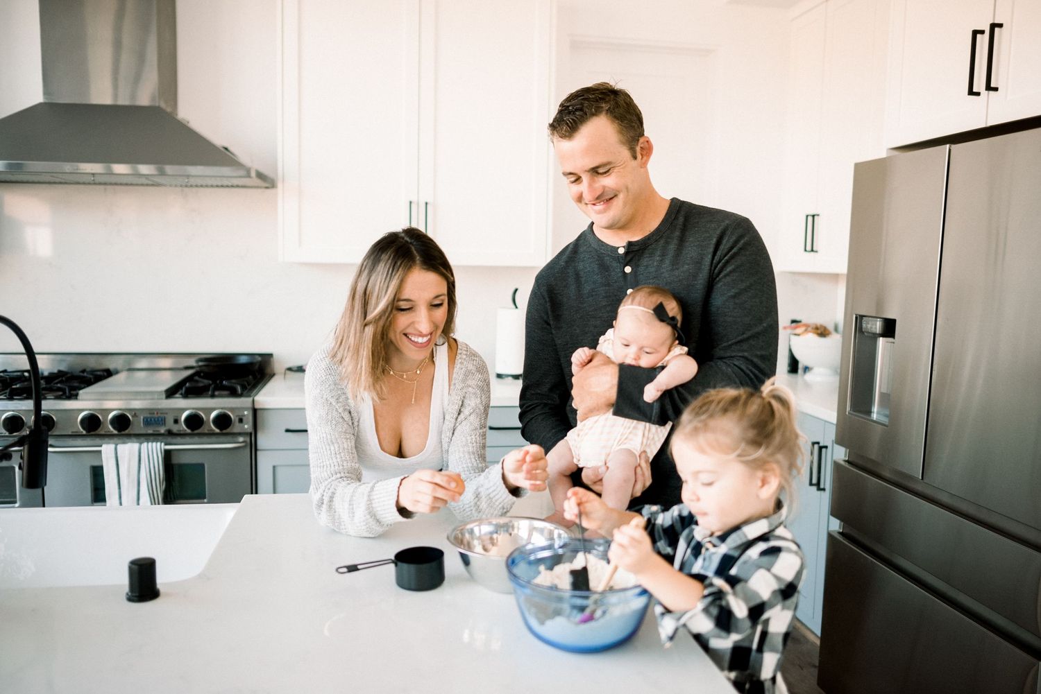 Happy family cooking together in modern white kitchen, parents with newborn baby and toddler son baking at counter.