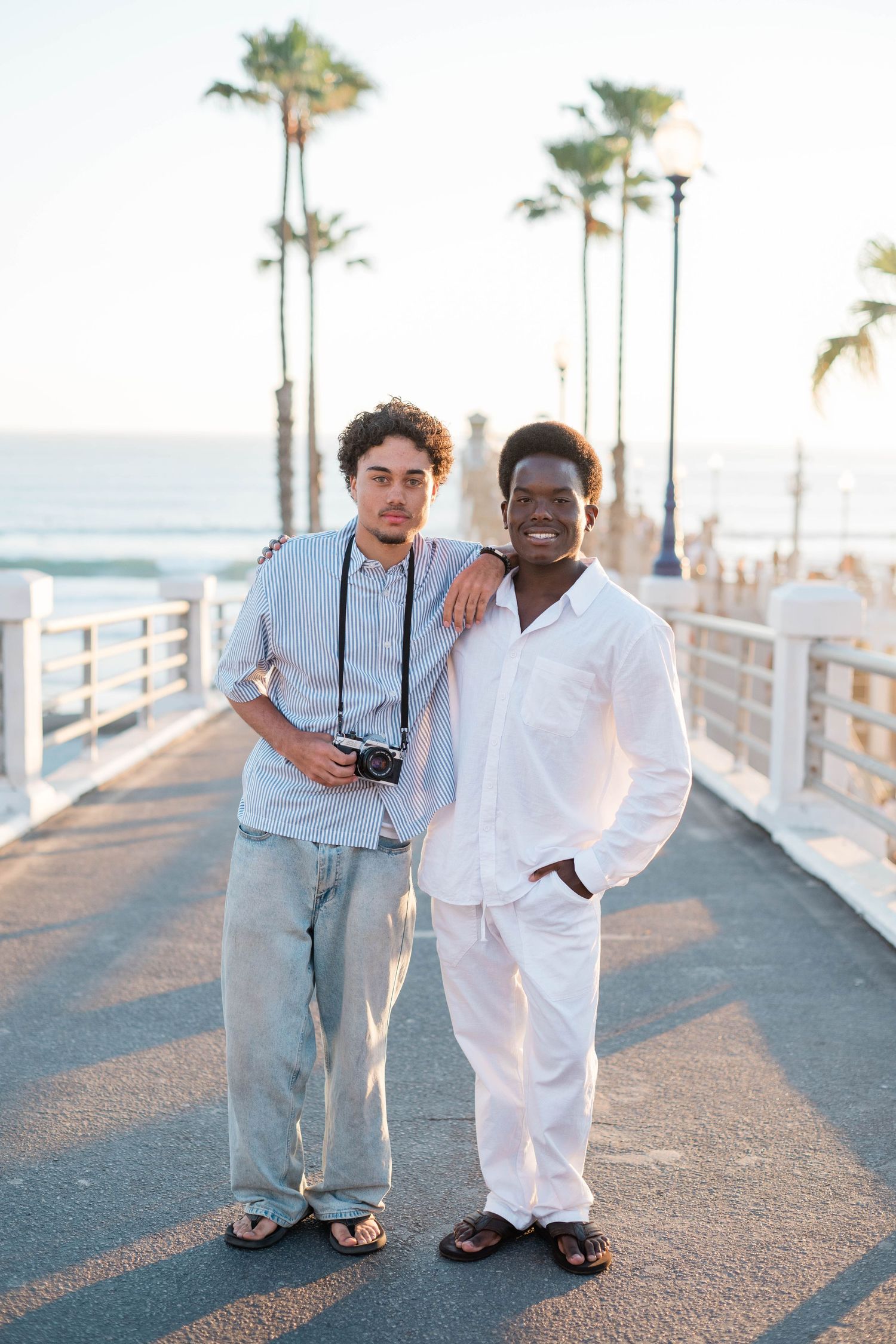 Two men posing on a sunny beachside pier with palm trees, one in white outfit and one in striped shirt holding a camera.