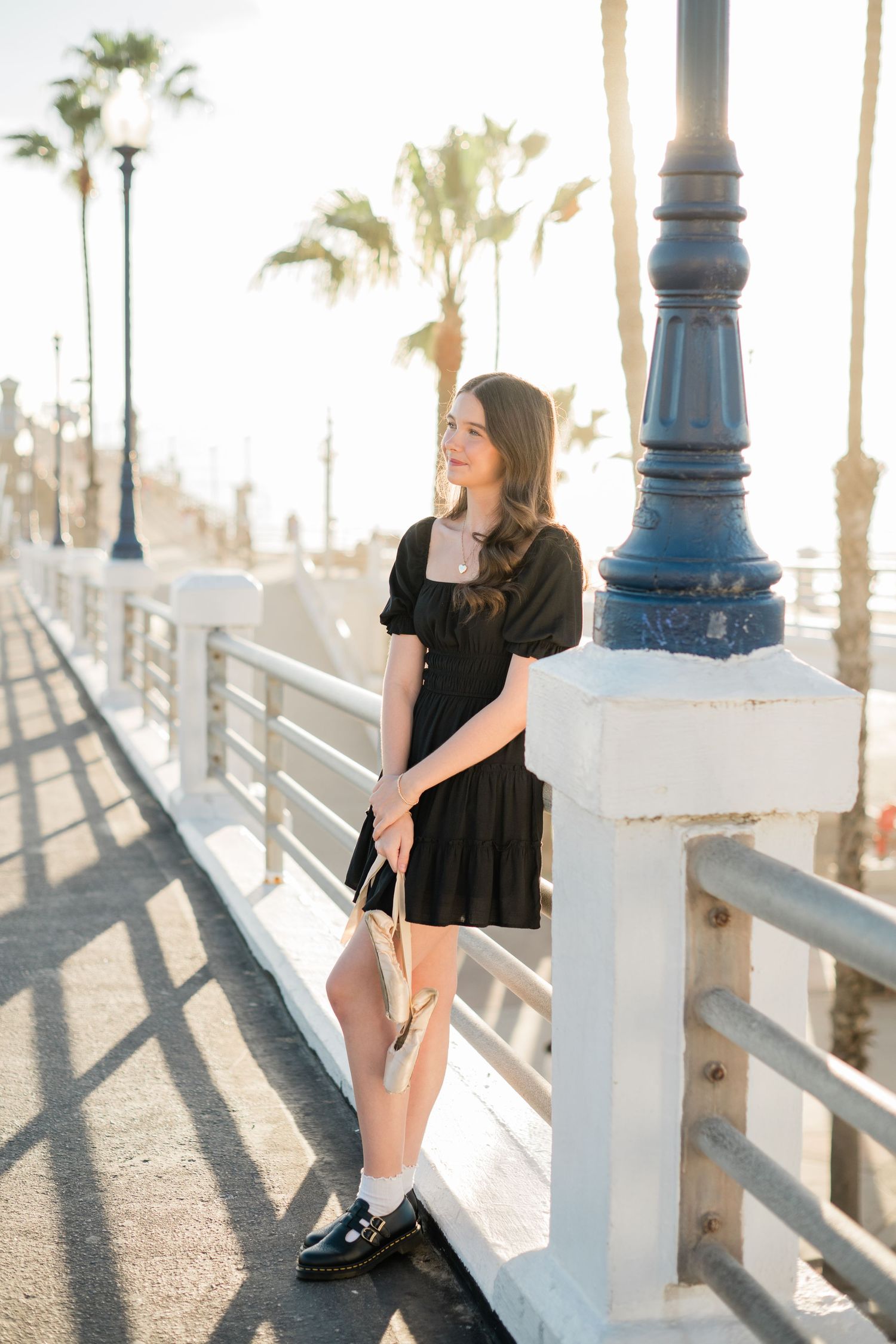 Young woman in black mini dress and loafers posing on a sunny boardwalk with palm trees in the background.