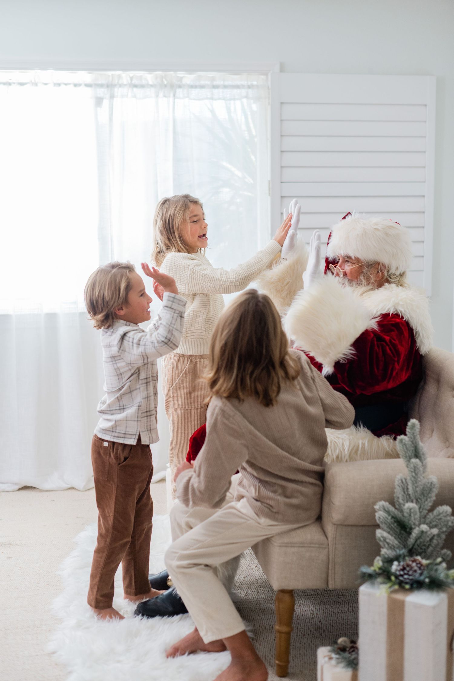 Three children give high fives to Santa Claus seated in a white armchair in a bright Christmas-decorated living room.