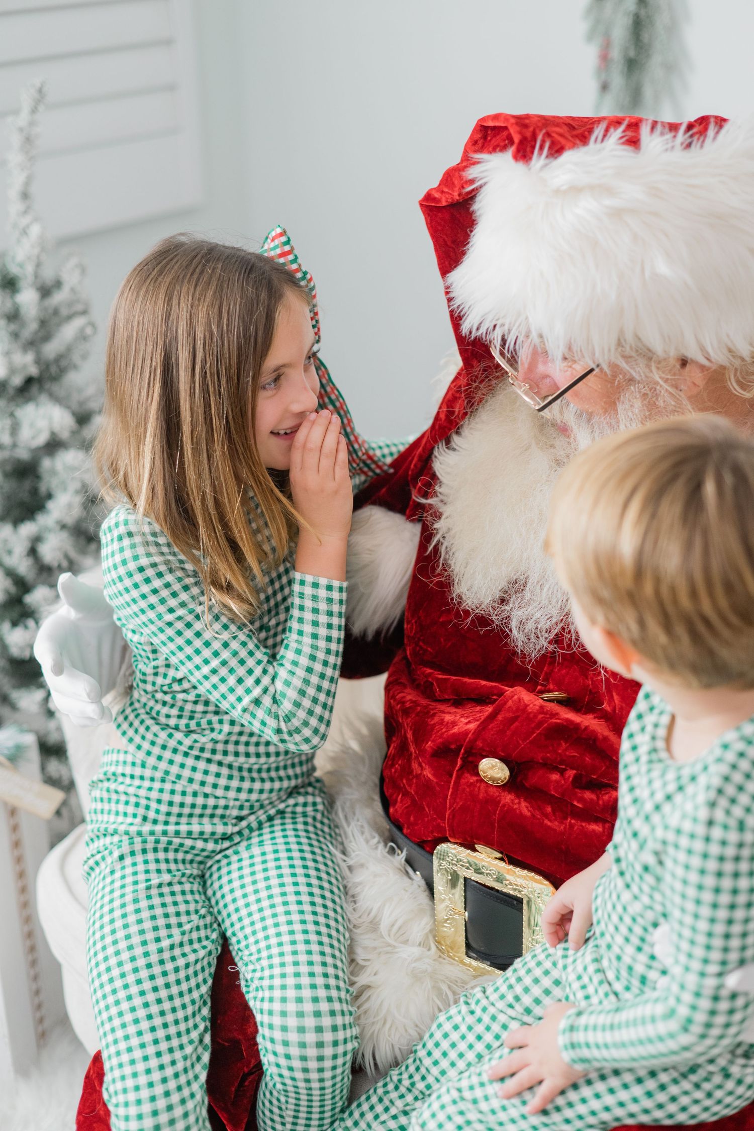 Young girl in green plaid pajamas whispers to Santa Claus while sitting on his lap during Christmas photo session.
