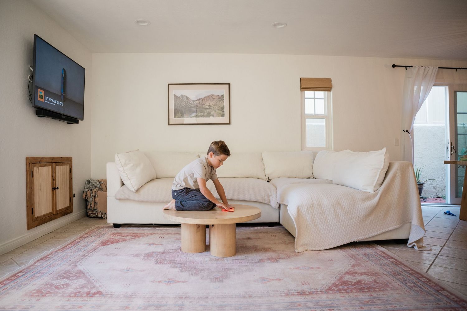 A young boy cleaning a round wooden coffee table in a bright, cozy living room with a white sectional sofa.