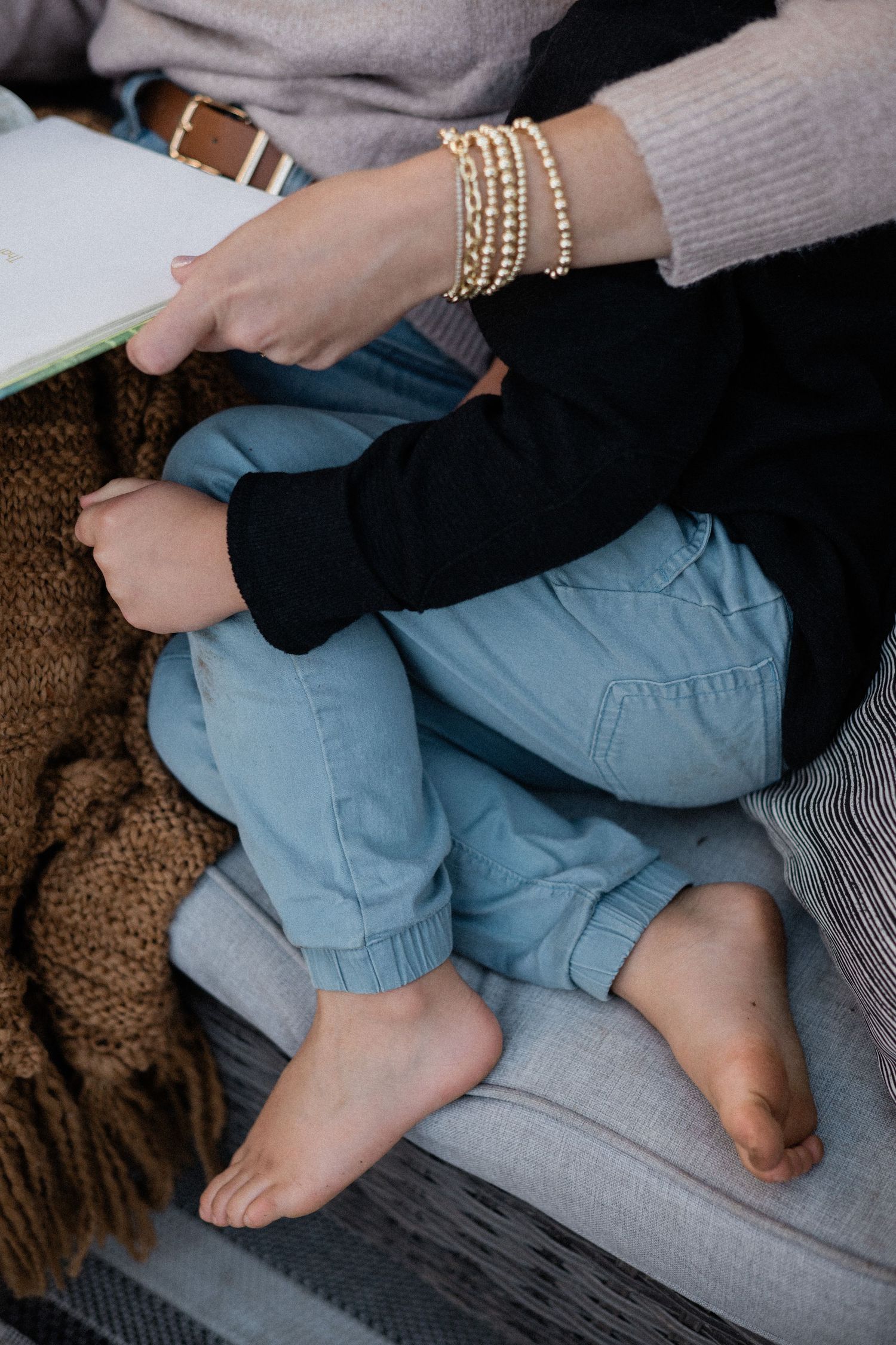 Close-up of a child sitting on a couch with a woman wearing gold bracelets holding an open book.