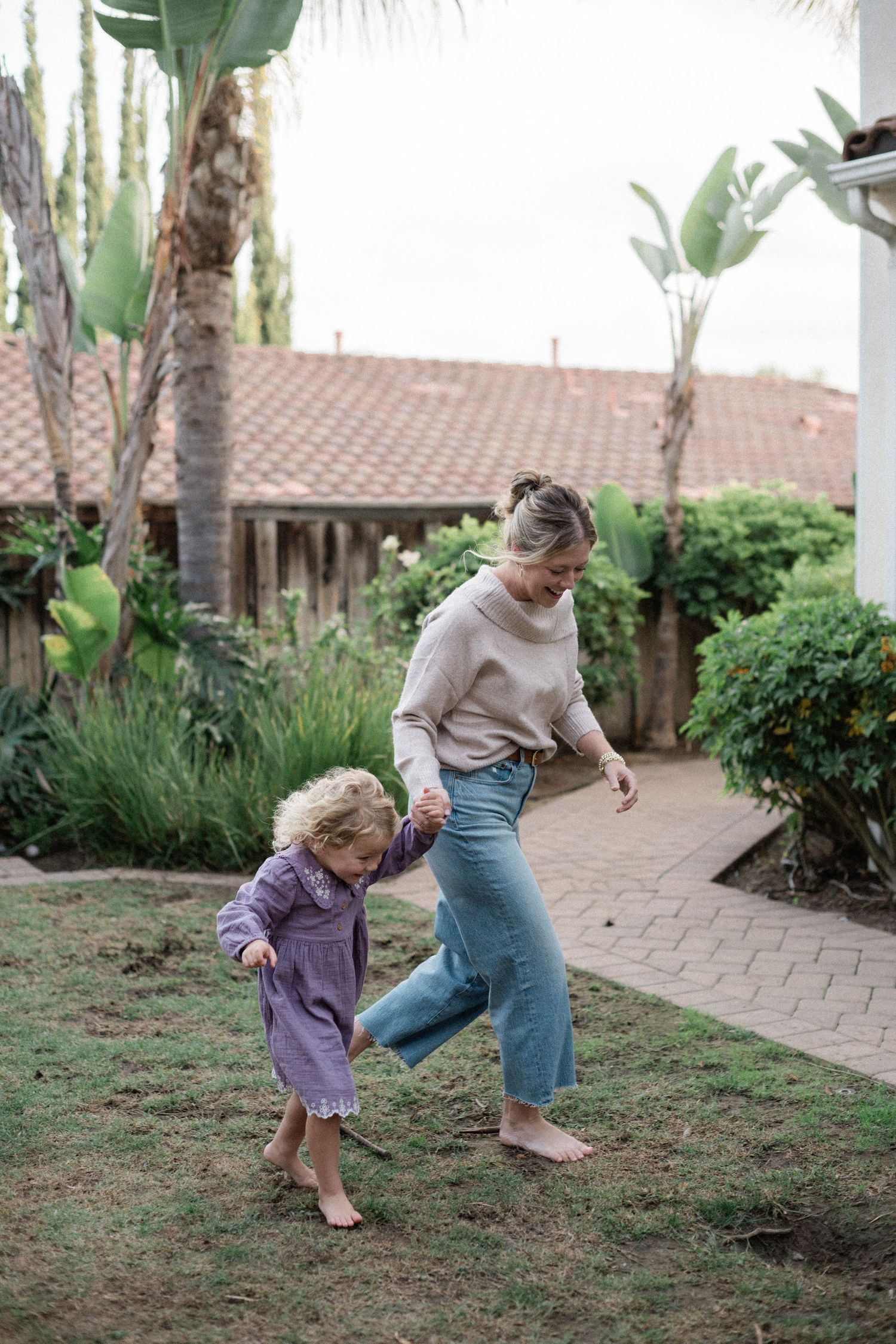 A mother and young child play barefoot on green grass in a lush backyard garden with tropical plants.