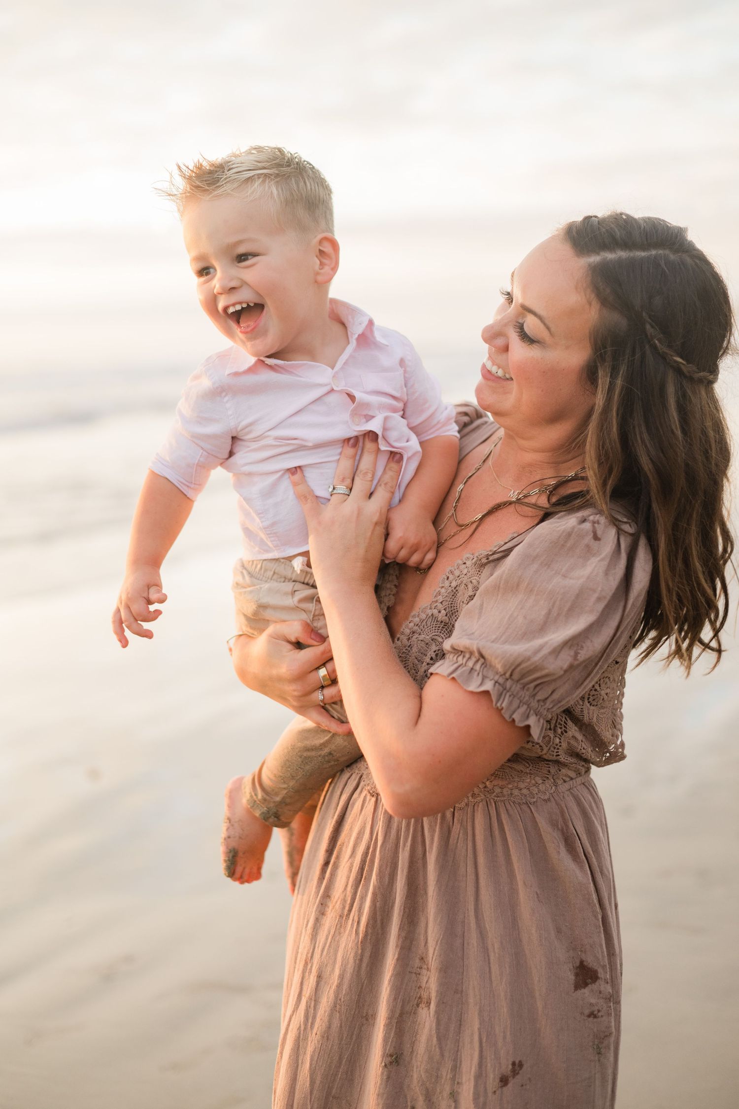 A joyful mother holds her laughing toddler boy on a sunny beach, both smiling in soft pastel outfits.