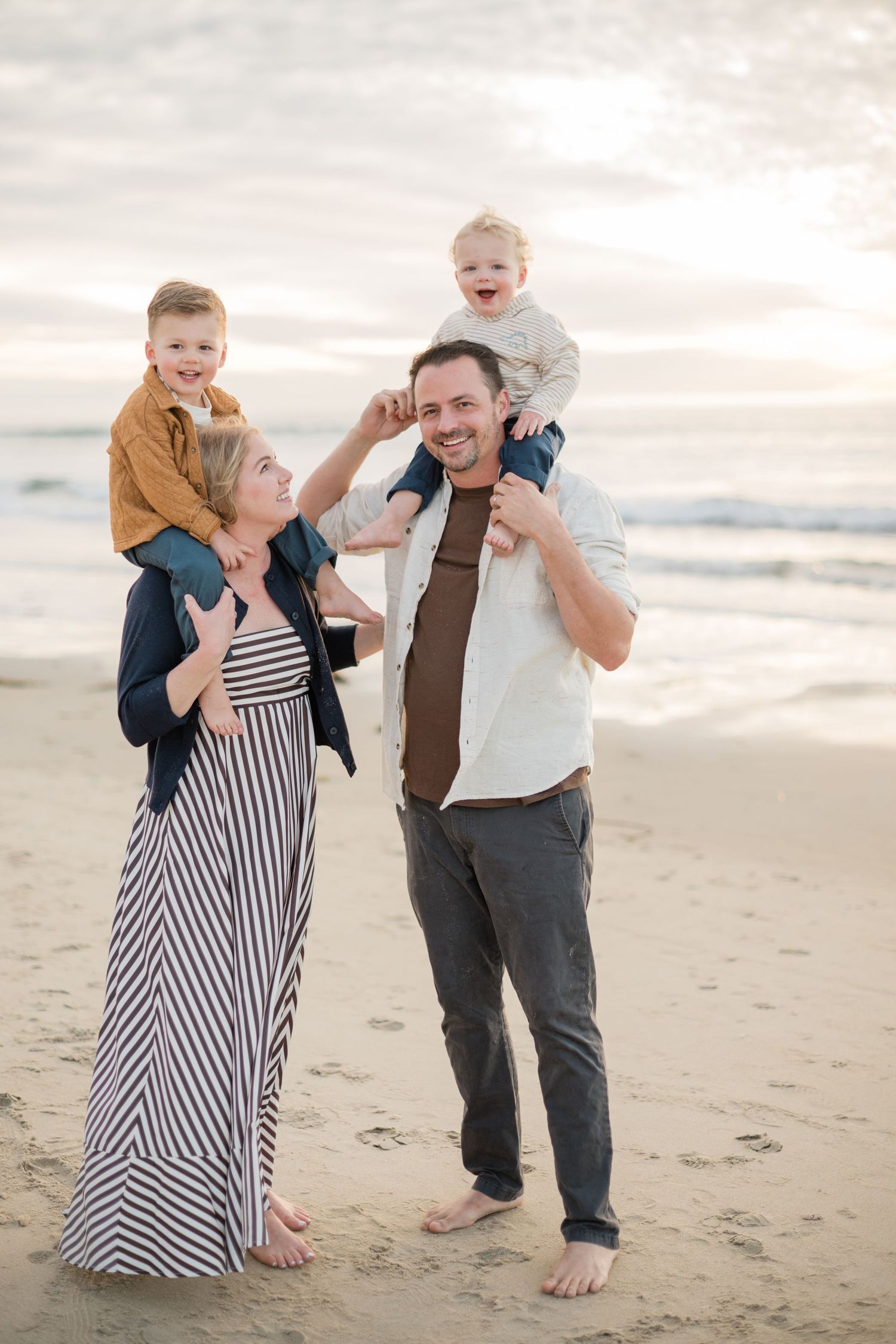 Happy family of four enjoying a beach sunset, parents carrying toddlers on shoulders, smiling and laughing together.