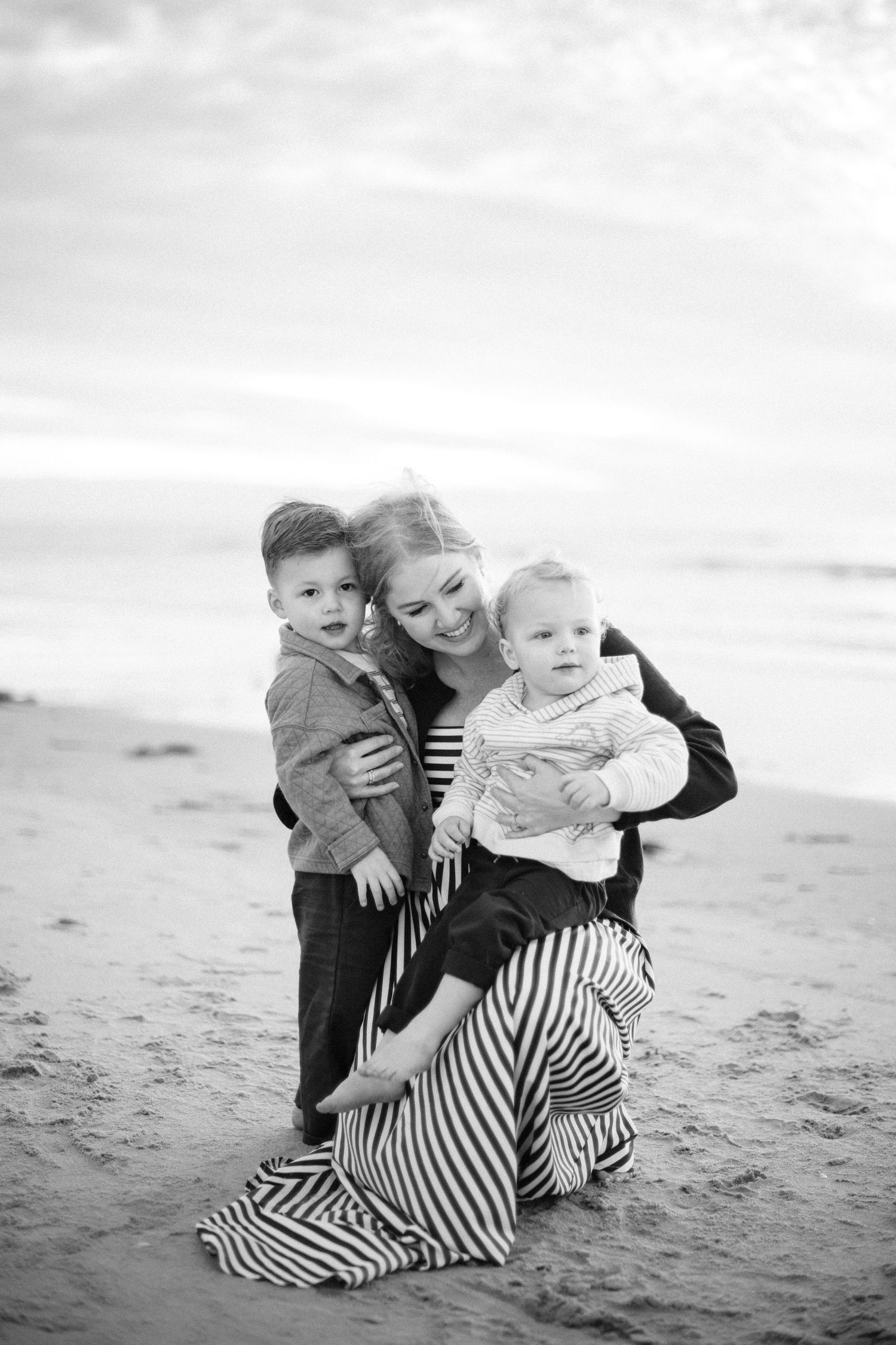 Woman in striped maxi skirt kneeling on beach hugging two young children in black and white photo.