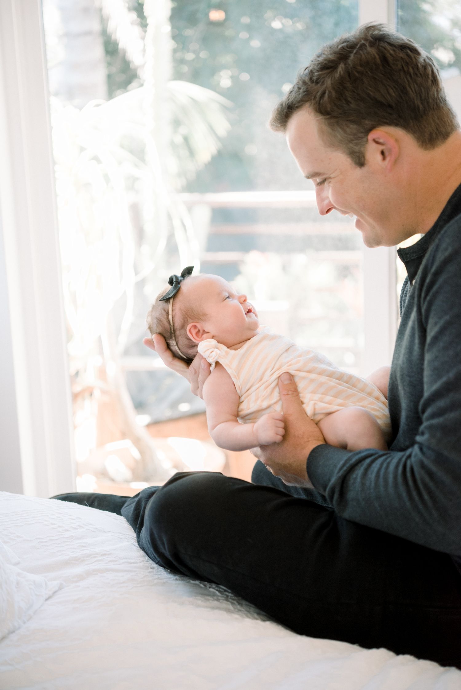 A father sits cross-legged on a bed, tenderly cradling his newborn baby near a bright window.