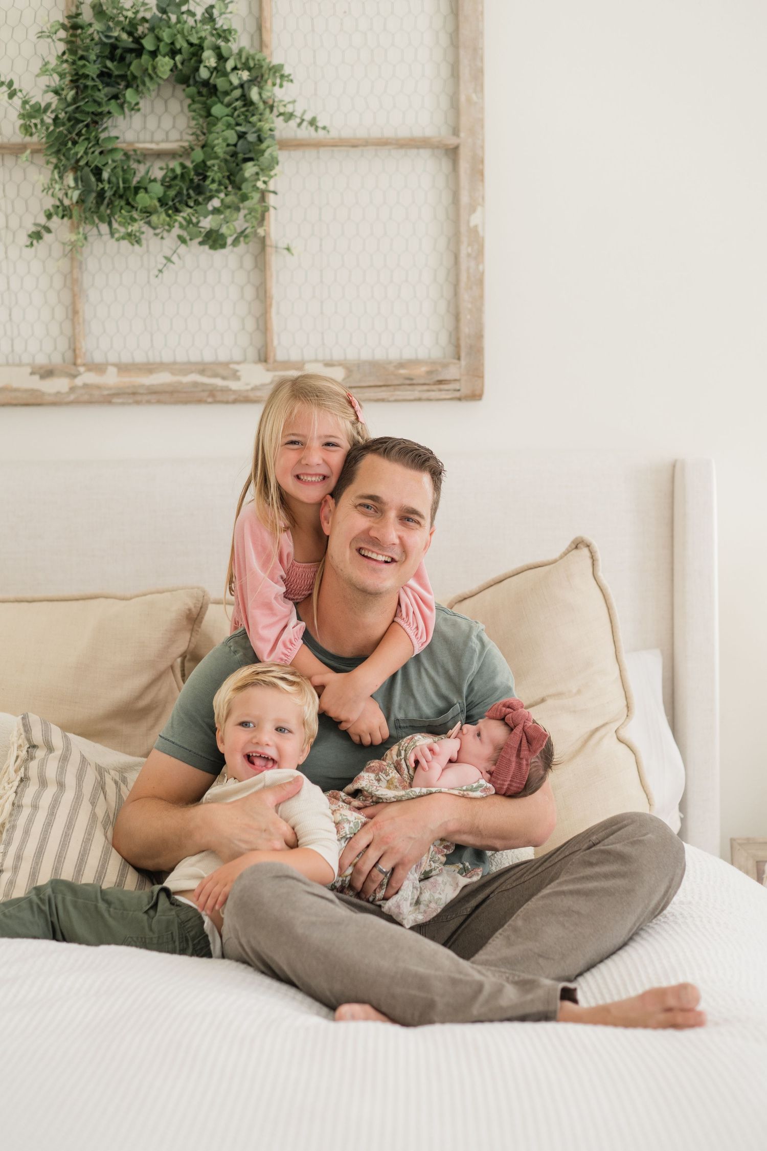 Smiling father sitting on white bed with three children including newborn baby girl and toddler son.