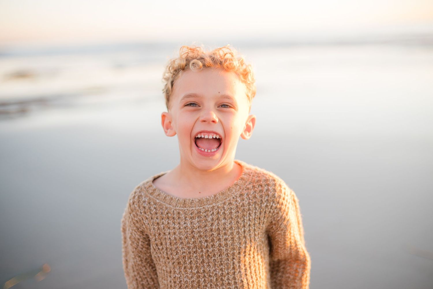 Happy young boy with curly hair laughing at the beach, wearing a tan knit sweater on a sunny day.