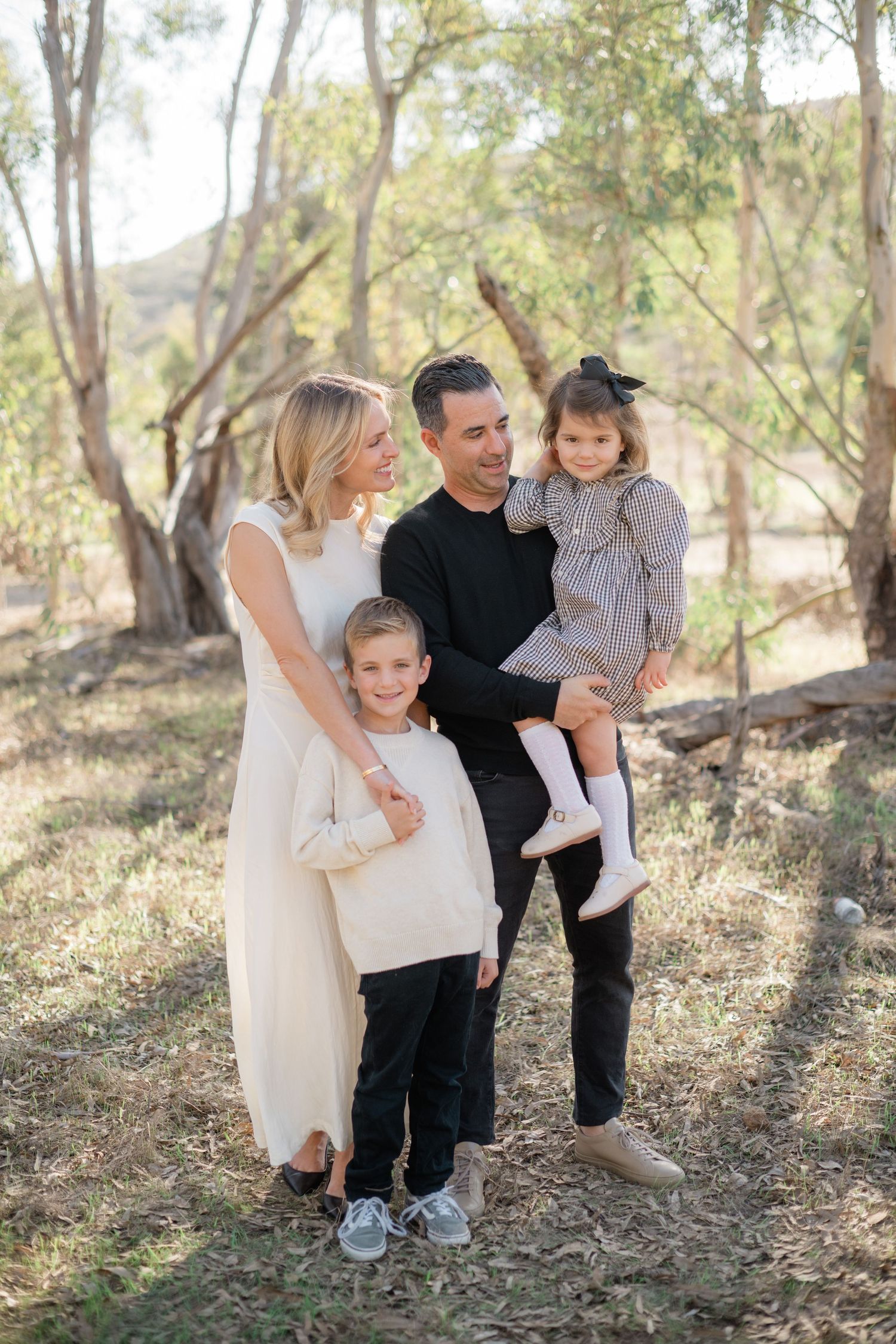 Family of four posing outdoors among eucalyptus trees, parents smiling at children in a warm natural setting.
