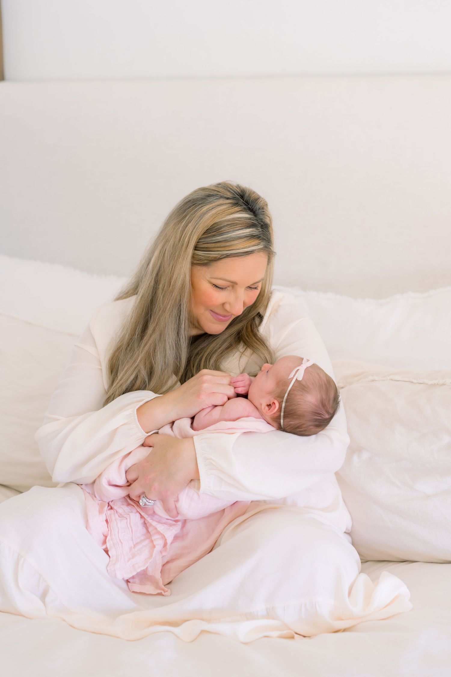 A mother with long blonde hair tenderly holds her sleeping newborn baby wrapped in pink on a white bed.