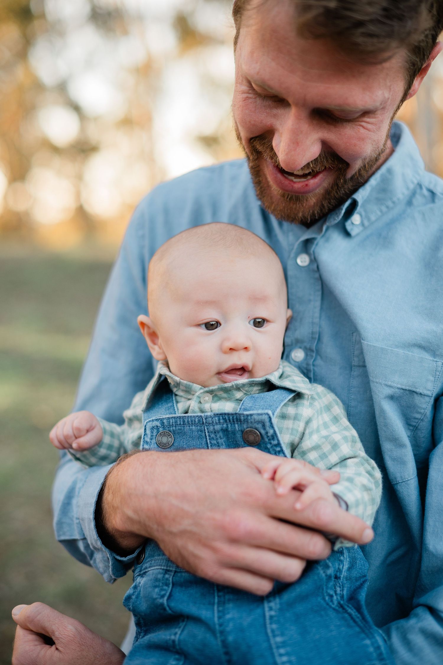 Smiling bearded father in blue shirt holding his baby boy dressed in plaid shirt and denim overalls outdoors.