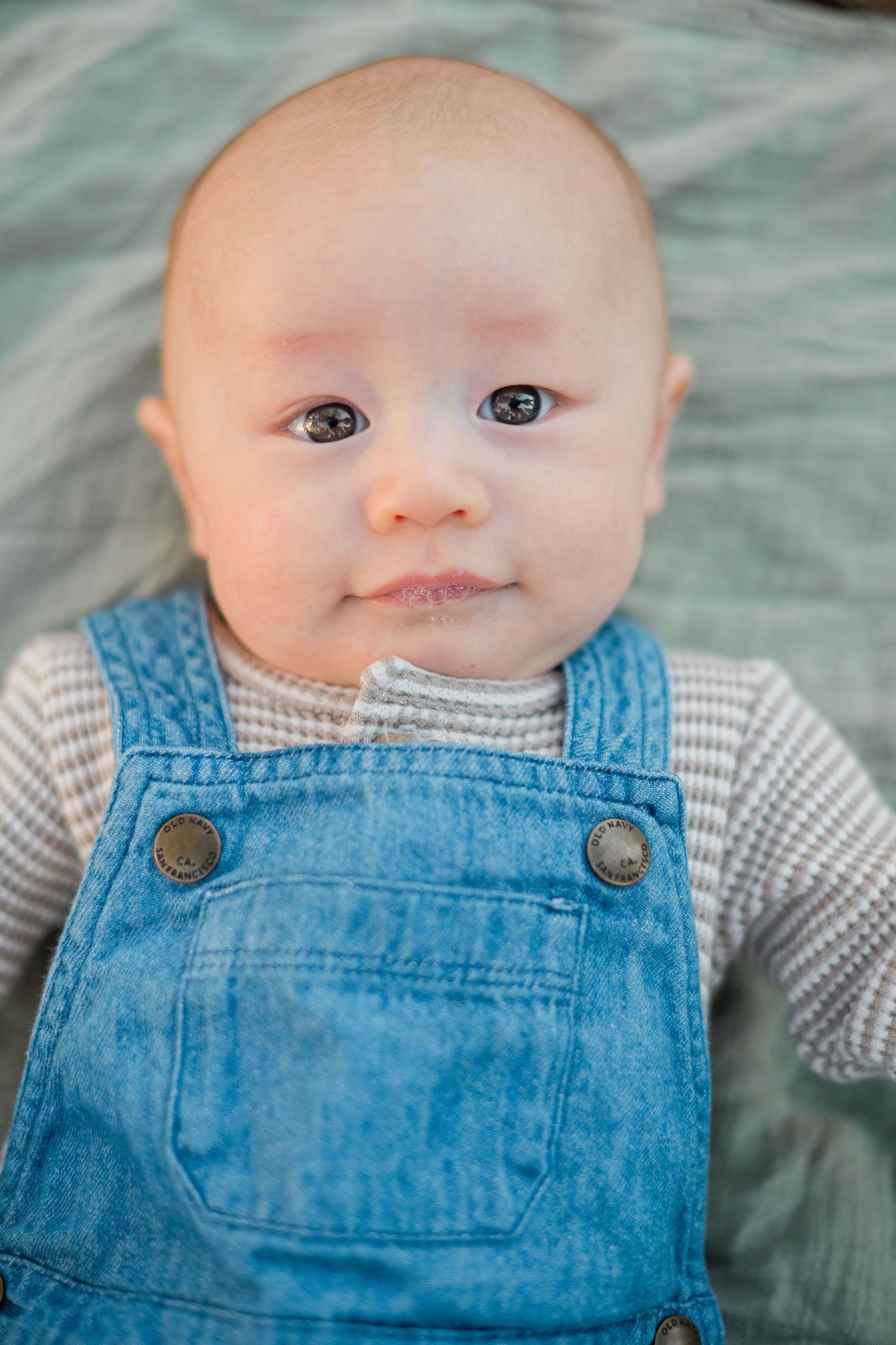 Adorable baby in denim overalls and striped shirt lying down, looking up with curious eyes.