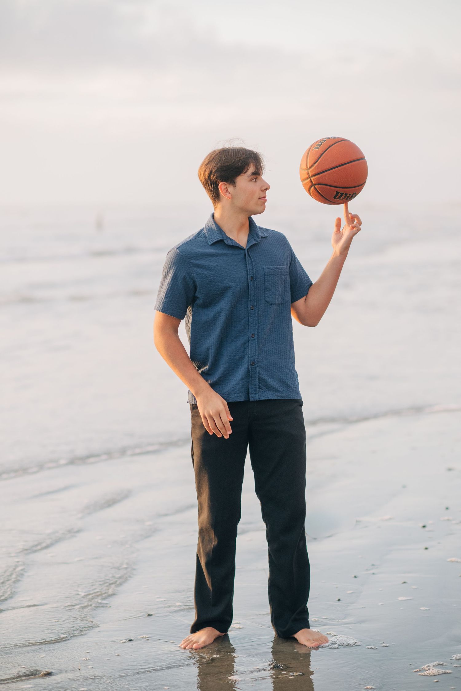 Young man spinning a basketball on his finger while standing barefoot on a beach shoreline.
