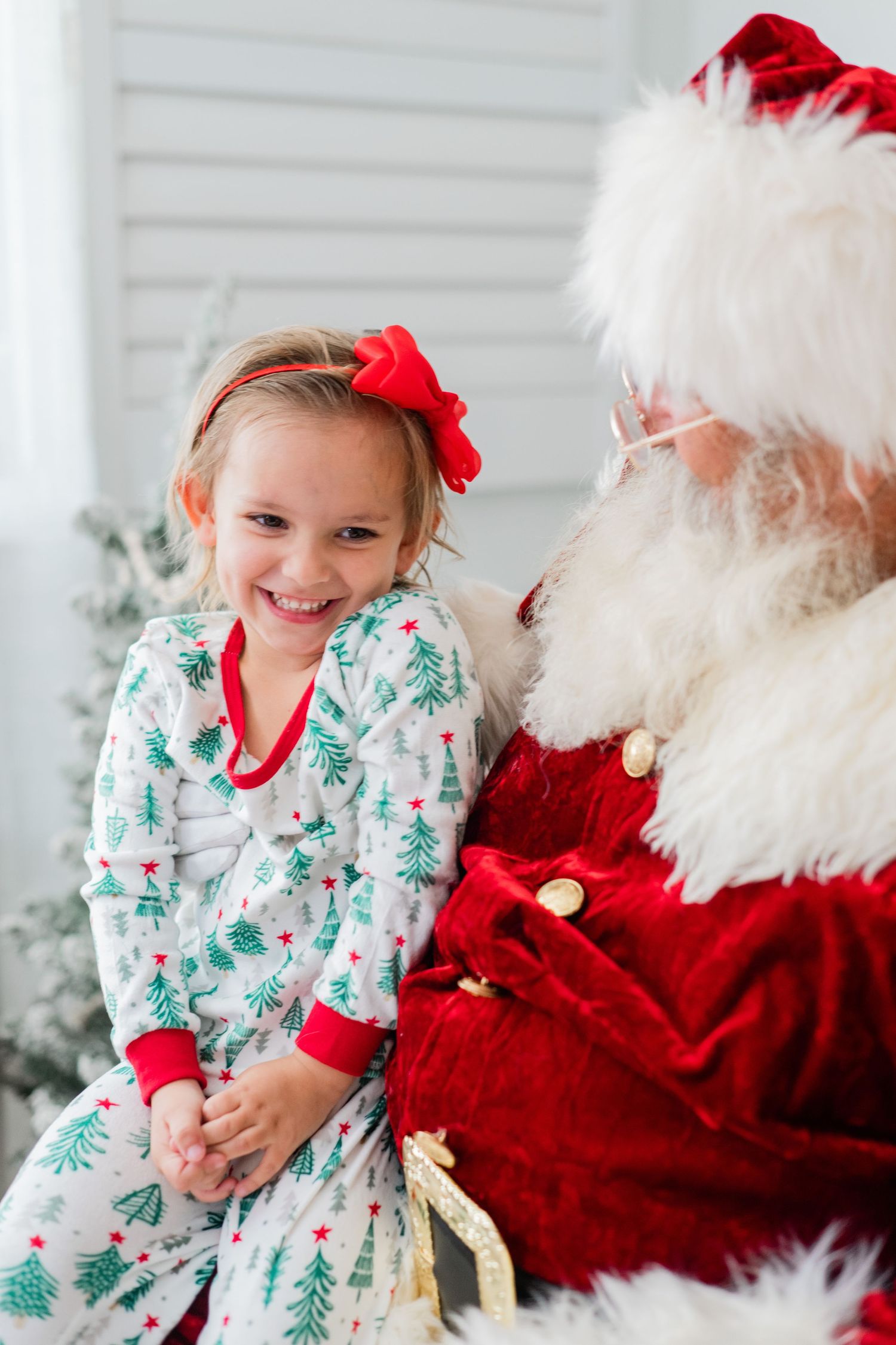 A smiling little girl in Christmas tree pajamas with a red bow sits happily on Santa Claus's lap.