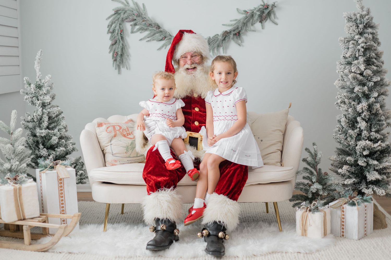 Two young girls sit on Santa Claus's lap on a cream sofa surrounded by snowy Christmas trees and gifts.