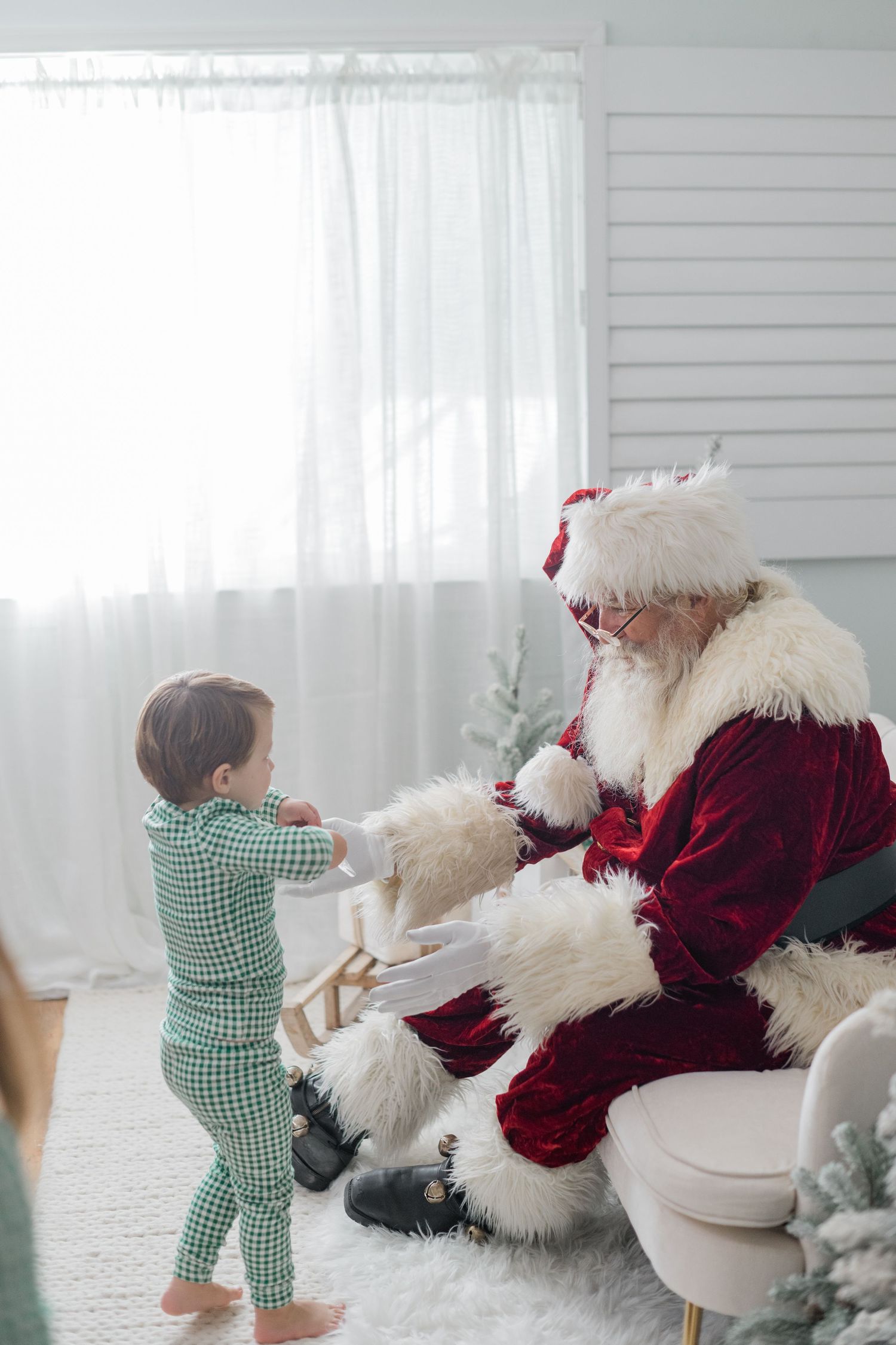 A toddler in green plaid pajamas hands a toy to Santa Claus in a bright white Christmas setting.