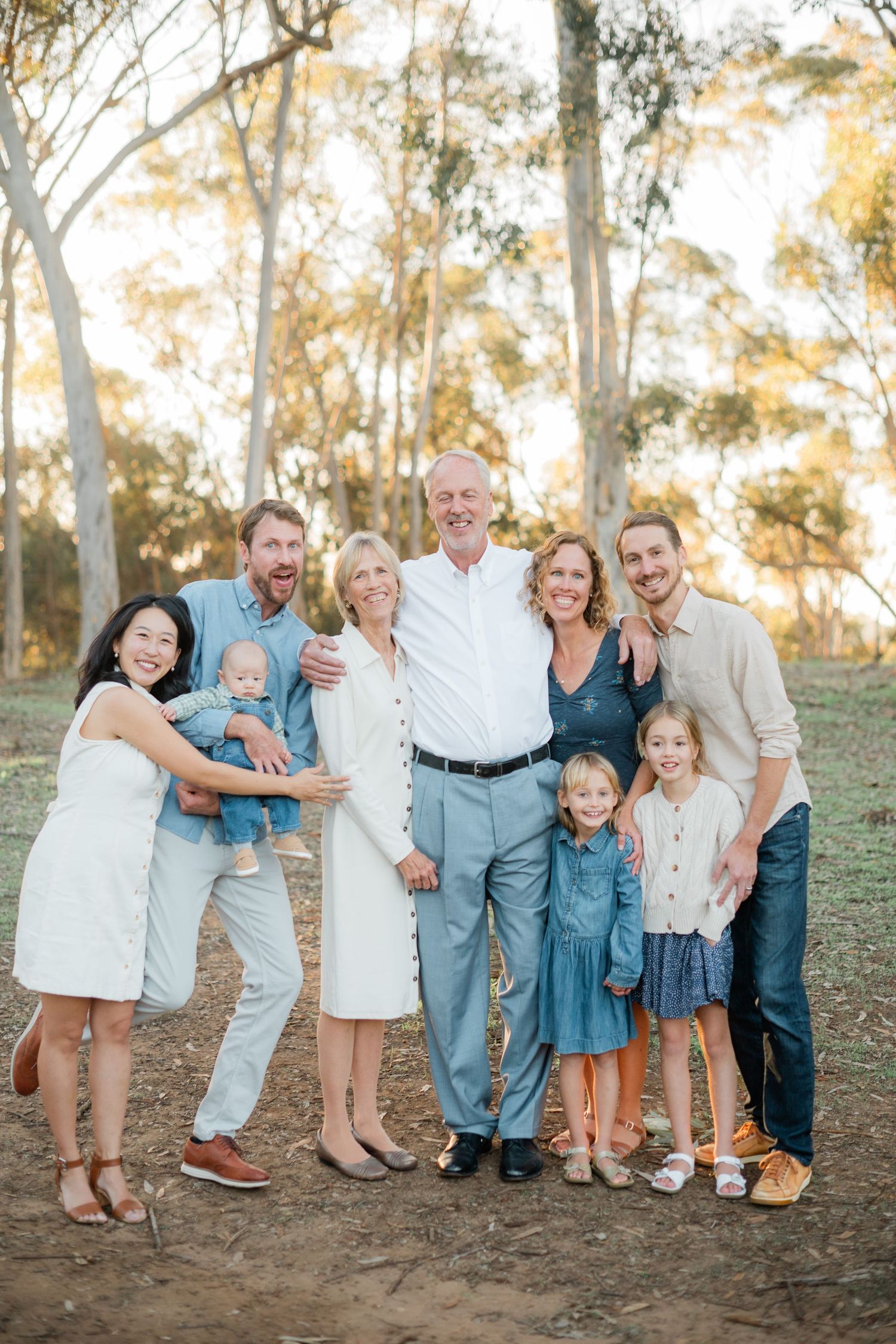 Multi-generational family portrait outdoors among eucalyptus trees, smiling together in coordinated casual outfits.